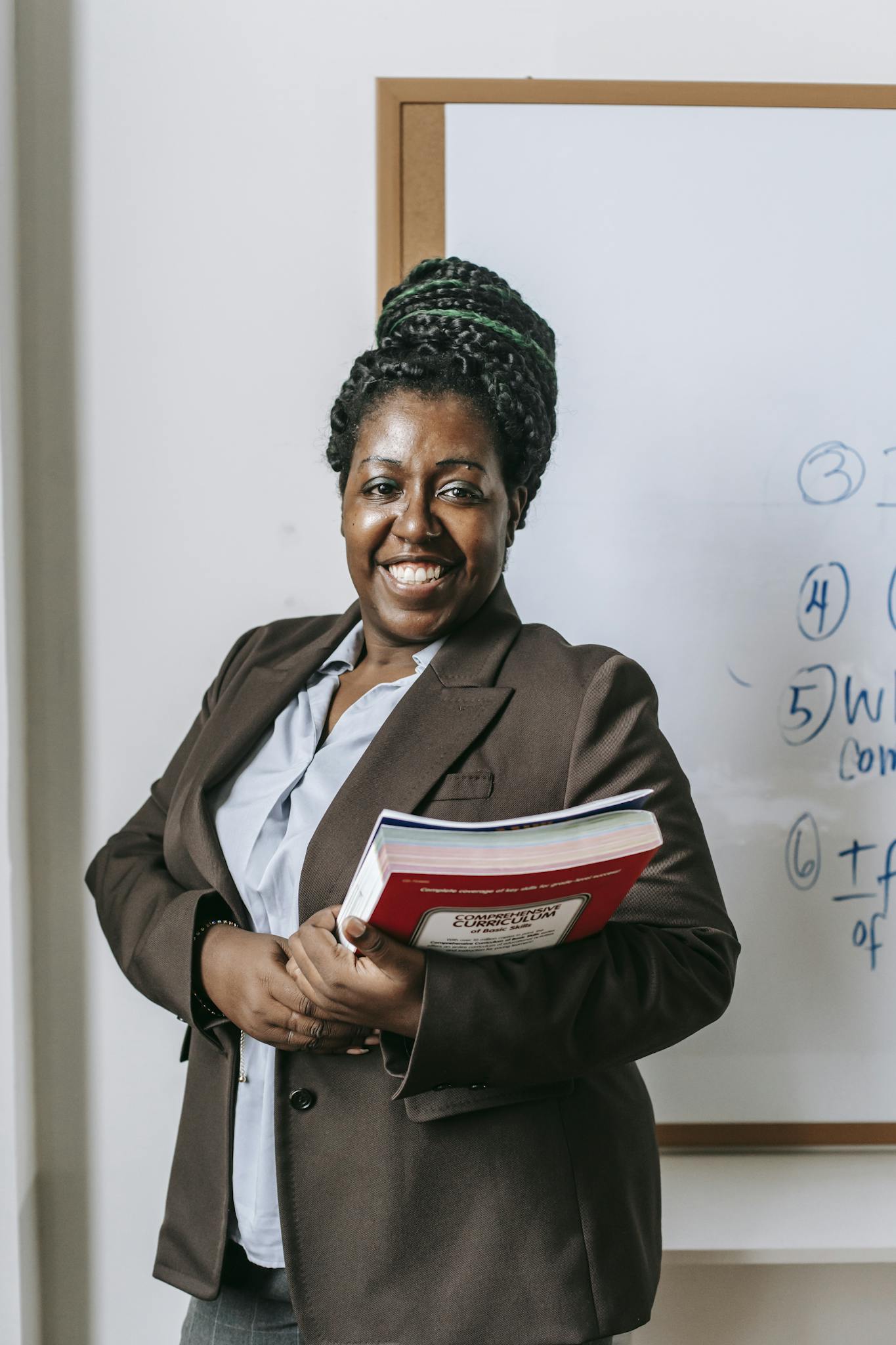 Smiling female lecturer holding books in front of classroom whiteboard.